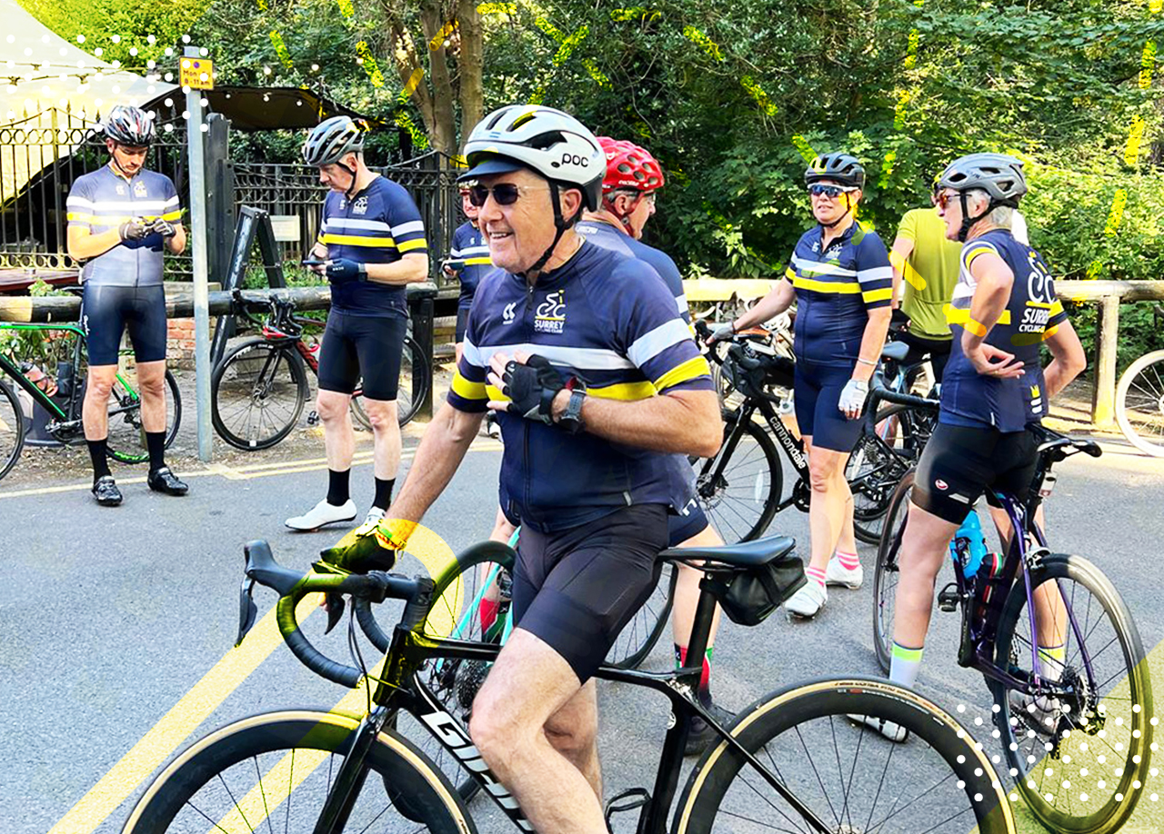 Group of cyclists in matching jerseys stand with their bikes on a road, chatting and preparing for a ride, surrounded by greenery.