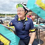 A man wearing cycling gear and a helmet sits at an outdoor table, smiling, with other people and tea cups in the background.