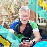 Older man in a green and blue cycling jersey sits outdoors at a table, smiling while looking at his smartphone. Colorful geometric overlays appear on the image.