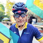A man wearing a cycling helmet, sunglasses, and a blue cycling jersey stands outdoors with colorful geometric shapes overlaid on the image.