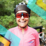 A person wearing a pink cycling jersey, bike helmet, and sunglasses smiles outdoors with greenery and a bicycle in the background.